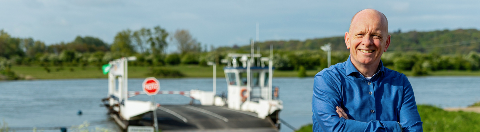 Arie kijkt naar de camera, armen over elkaar, voor een rivier met een veerpont.